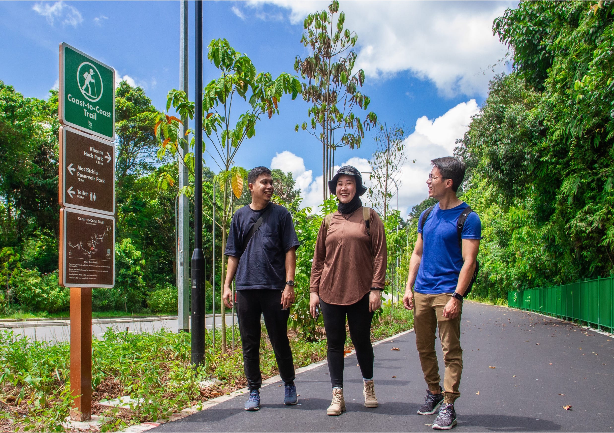 Three people stand on a sunny trail near a green Coast-to-Coast Trail signpost surrounded by lush trees and bushes.